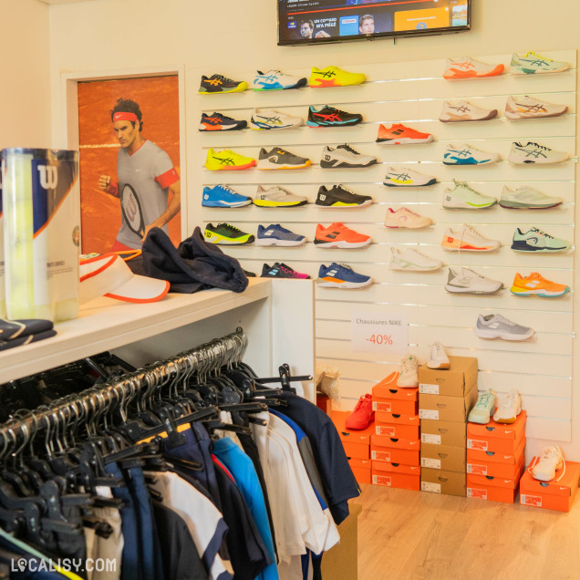 Des chaussures de tennis exposées sur un mur, des vêtements sur un portant, et plusieurs boîtes de chaussures orange au magasin “Tennispro Nandrin” à Nandrin.