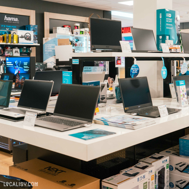 Plusieurs ordinateurs portables exposés sur une table centrale, avec leurs écrans ouverts chez “DGS Informatique” à Saint-Georges-sur-Meuse. Le magasin est bien éclairé et organisé, avec des produits emballés sur les étagères en arrière-plan et des matériaux promotionnels visibles.