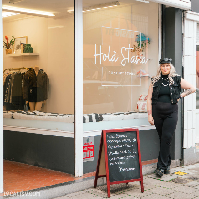 Devanture du magasin "Holà Stasia" à Jemeppe, avec une personne qui se tient devant le magasin. La vitrine expose des vêtements et il y a un panneau sandwich devant avec des inscriptions.