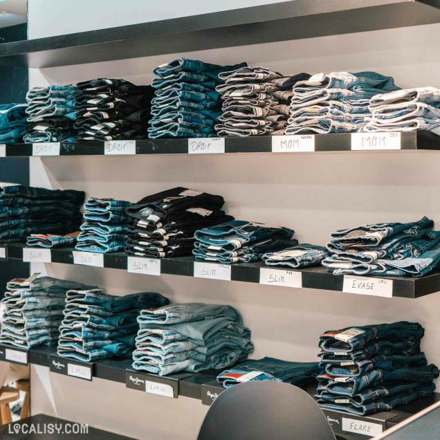 Rayons de magasin ''GOMINA'' à Liège avec jeans, t-shirts et casquettes, incluant des articles Replay.