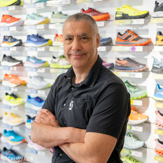 Une personne, probablement le gérant du magasin d'articles de course à pied Xrun à Battice, debout devant un présentoir mural de chaussures de course colorées.