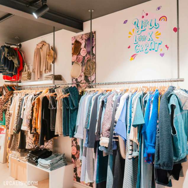 Des vêtements suspendus sur des cintres le long de deux tringles parallèles dans le magasin de vêtements Char'li Concept à Malmedy. Les vêtements sont variés, comprenant des jeans, des vestes, des chemises et des pulls de différentes couleurs et motifs. Au-dessus des tringles, il y a des étagères avec des accessoires comme des sacs à main et des chapeaux.
