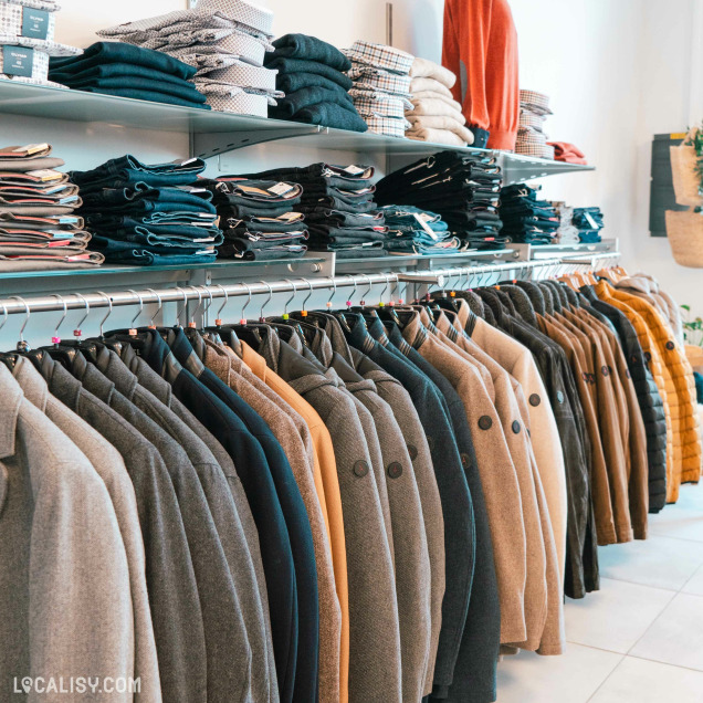 Des étagères remplies de vêtements pliés, principalement des pantalons et des chemises, ainsi qu'une rangée de manteaux suspendus sur des cintres dans le magasin de vêtements pour hommes De Fil En Costume à Chaudfontaine.
