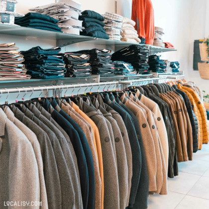 Des étagères remplies de vêtements pliés, principalement des pantalons et des chemises, ainsi qu'une rangée de manteaux suspendus sur des cintres dans le magasin de vêtements pour hommes De Fil En Costume à Chaudfontaine.