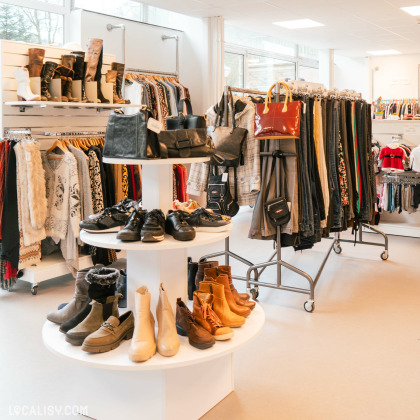 Des étagères et des portants remplis de vêtements variés, tels que des manteaux, des vestes, des pantalons et des pulls dans le magasin de vêtements Rebelote à Comblain-au-Pont. Au centre, il y a un présentoir circulaire à deux niveaux avec des chaussures et des sacs à main. L'ambiance du magasin est lumineuse et bien organisée.