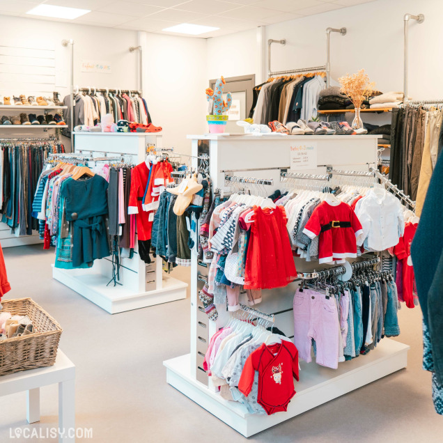 Une variété d'étagères et portants remplis de vêtements de différentes tailles et styles, y compris des vêtements pour bébés et enfants dans le magasin de vêtements Rebelote à Comblain-au-Pont. On peut voir des vêtements suspendus sur des cintres, des chaussures sur des étagères, et des accessoires comme des chapeaux.