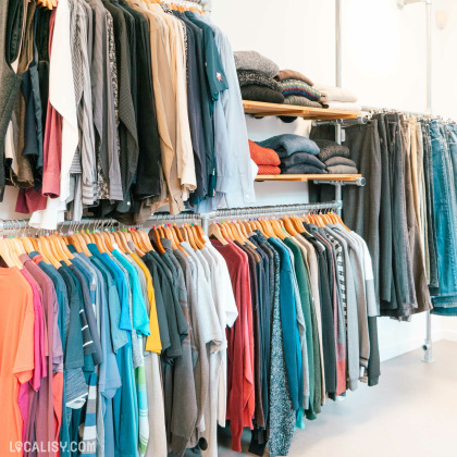 L'intérieur du magasin de vêtements Rebelote à Comblain-au-Pont, avec des étagères et des portants remplis de divers articles. En haut, il y a des chemises et des pantalons suspendus, ainsi que des piles de pulls et de vêtements pliés sur des étagères. En bas, il y a une rangée de t-shirts et de pulls de différentes couleurs accrochés à des cintres.