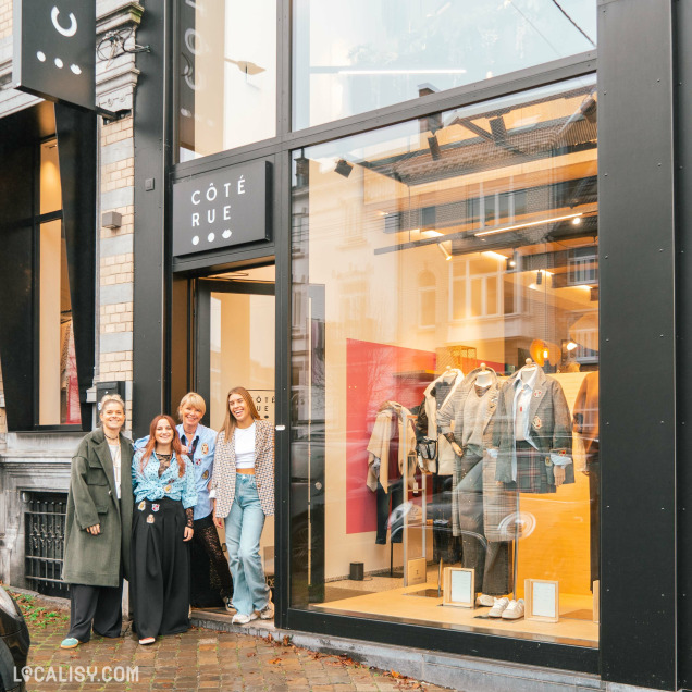 Devanture du magasin d'accessoires de mode Côté Rue à Heusy. La façade est moderne avec de grandes vitrines exposant des vêtements. Devant l'entrée, quatre personnes posent pour une photo. Le trottoir pavé est humide, suggérant une pluie récente. Le magasin semble être une boutique de mode, mettant en avant des vêtements et des accessoires tendance.