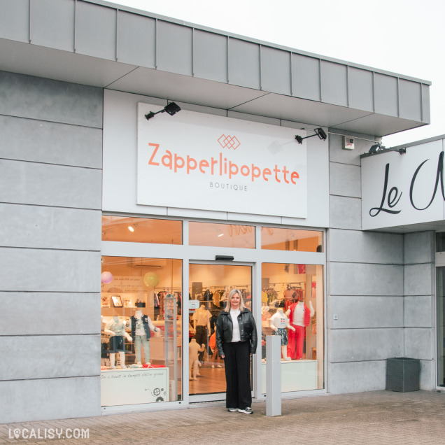 Façade du magasin Zapperlipopette à Herve, avec une femme souriante devant l’entrée. Le logo du magasin est bien visible au-dessus de la porte vitrée, à côté d’un autre commerce.