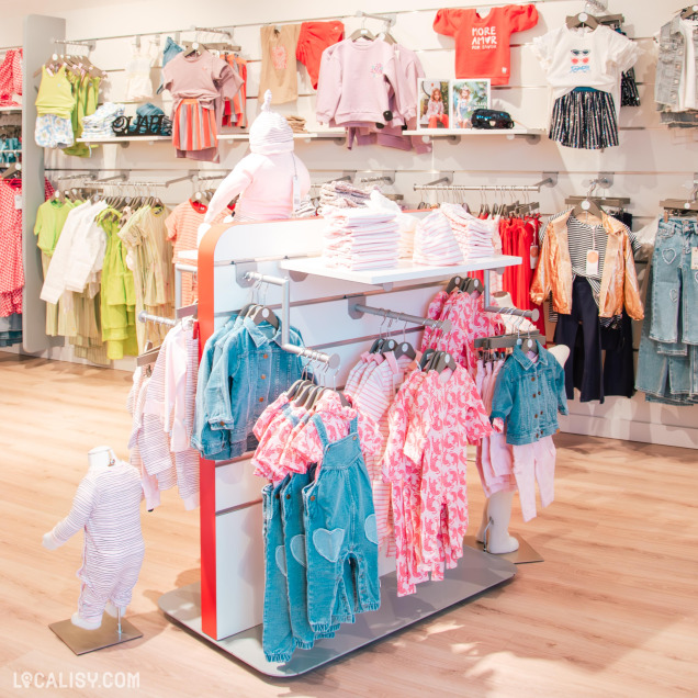 Présentation de vêtements pour bébés et jeunes enfants dans des tons pastel et colorés, avec des mannequins miniatures et des portants bien organisés.