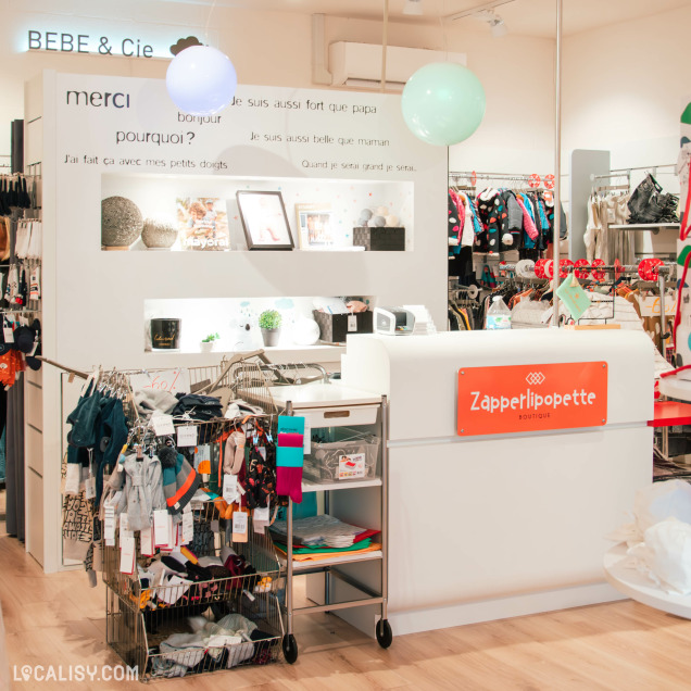 Vue intérieure du magasin Zapperlipopette, avec un comptoir blanc arborant le logo du magasin en orange. À l’arrière, un mur décoré de phrases inspirantes et une section d’articles pour bébés et enfants.
