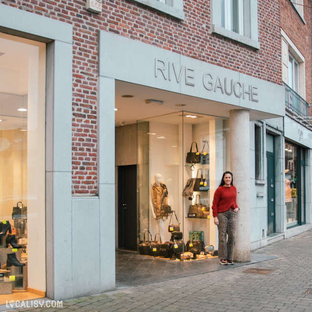 La façade du magasin "Rive Gauche", une boutique de mode chic avec une devanture en briques et une entrée moderne en verre. Une femme souriante, portant un pull rouge et un pantalon imprimé, pose devant la boutique