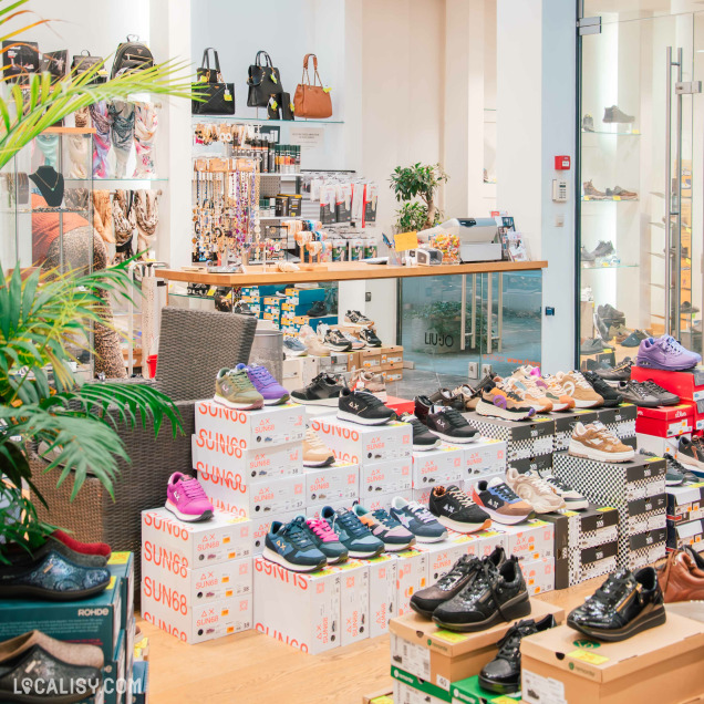 Un magasin de mode et de chaussures proposant une large sélection de baskets de différentes marques, exposées sur des boîtes empilées. En arrière-plan, on aperçoit des sacs à main, des accessoires et un comptoir de caisse avec divers produits.