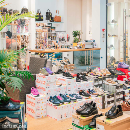 Un magasin de mode et de chaussures proposant une large sélection de baskets de différentes marques, exposées sur des boîtes empilées. En arrière-plan, on aperçoit des sacs à main, des accessoires et un comptoir de caisse avec divers produits.