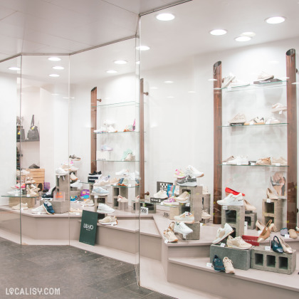 Un coin d’exposition du magasin, où des sneakers et chaussures élégantes sont soigneusement disposées sur des étagères en verre et des supports en béton, sous un éclairage moderne.