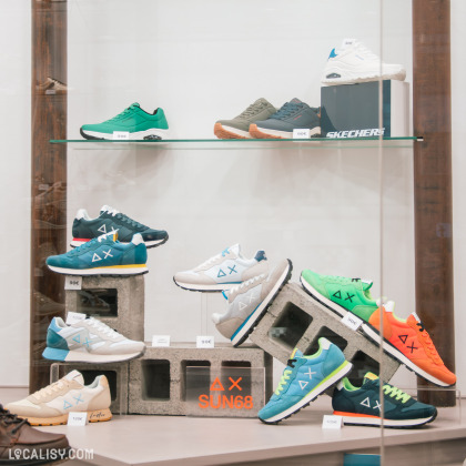 Gros plan sur une vitrine du magasin, mettant en avant une collection de baskets de marques comme SUN68 et Skechers. Les chaussures sont exposées sur des étagères et des blocs de béton, avec des étiquettes de prix bien visibles.