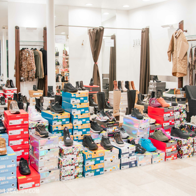 Vue intérieure de la boutique, avec un grand choix de chaussures et bottes disposées sur des piles de boîtes colorées. En arrière-plan, des cabines d’essayage avec rideaux et des portants de vêtements d’hiver complètent l’agencement du magasin.