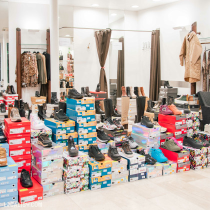 Vue intérieure de la boutique, avec un grand choix de chaussures et bottes disposées sur des piles de boîtes colorées. En arrière-plan, des cabines d’essayage avec rideaux et des portants de vêtements d’hiver complètent l’agencement du magasin.