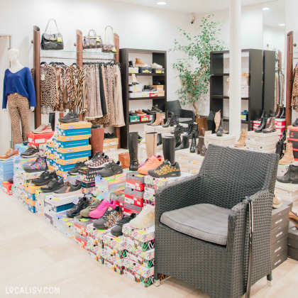 Intérieur d’une boutique de mode et de chaussures, présentant un large choix de baskets colorées et de bottes exposées sur des boîtes empilées. En arrière-plan, des portants avec des vêtements à imprimés léopard et une étagère de chaussures sont visibles, accompagnés d’un mannequin habillé.