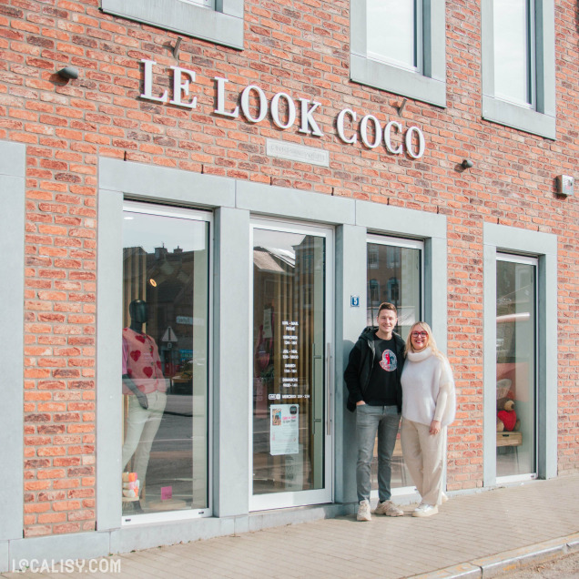 Façade du magasin "Le Look Coco" : Une boutique en briques rouges avec une enseigne en lettres blanches affichant "LE LOOK COCO". Deux personnes souriantes, un homme en sweat noir et une femme en pull blanc, posent devant l'entrée vitrée du magasin. À travers les vitrines, on peut apercevoir un mannequin habillé et des articles de mode.