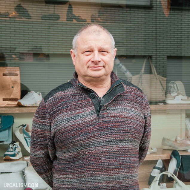 Un homme au visage souriant, vêtu d'un pull à motifs, pose devant la vitrine du magasin "Chaussures Lehane". En arrière-plan, des sacs et des chaussures sont exposés derrière la vitre.