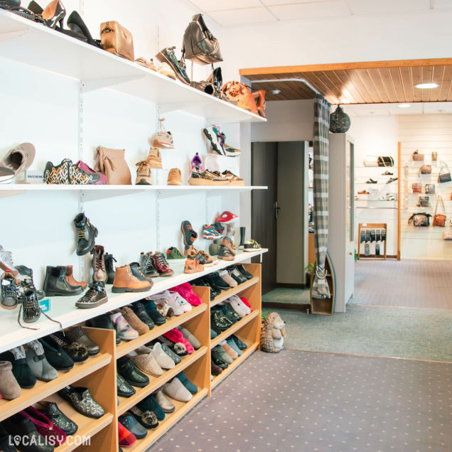 Autre vue de l’intérieur du magasin, montrant un large choix de chaussures et accessoires. L'agencement est clair et organisé, avec des chaussures alignées sur des étagères blanches et des meubles en bois, créant une ambiance chaleureuse et accueillante.
