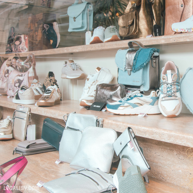 Une vitrine de magasin de chaussures et maroquinerie mettant en avant des sacs à main, des baskets et des accessoires aux tons pastel et métallisés. Les articles sont soigneusement disposés sur des étagères en marbre beige.
