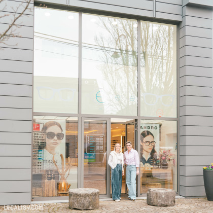 Devanture d'un magasin d'optique moderne avec une grande baie vitrée. Deux femmes souriantes se tiennent devant l'entrée, accueillant les clients. La vitrine affiche des publicités de lunettes de marque et des horaires d'ouverture.