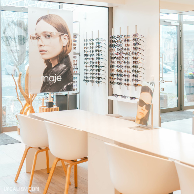 Un espace d’accueil élégant dans un magasin d’optique, avec une table de consultation blanche et des chaises design. Des présentoirs de lunettes sont visibles en arrière-plan, accompagnés d’affiches de marques connues.
