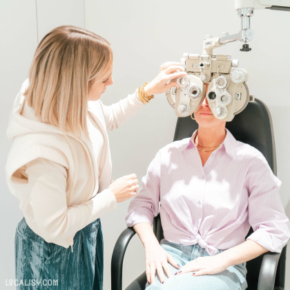 Une opticienne réalise un test de vue sur une patiente à l'aide d'un phoroptère. La patiente est assise dans un fauteuil noir, portant une chemise rose clair et un jean, tandis que l'opticienne ajuste l'appareil avec précision.