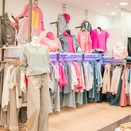 Intérieur d’une boutique de prêt-à-porter avec une sélection de vêtements dans les tons rose et denim. Mannequins habillés en tenues tendance, avec des sweats, vestes et jeans. Ambiance lumineuse et agencement soigné.