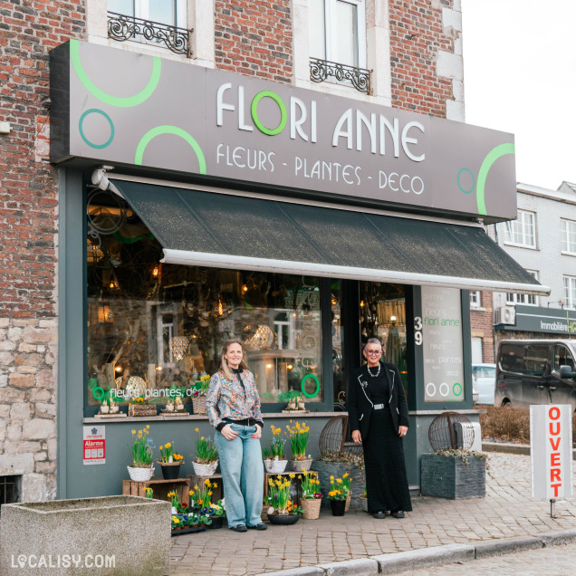 Une boutique de fleurs nommée "Flori Anne" avec une façade en briques rouges et une enseigne grise ornée de cercles verts. Deux femmes souriantes posent devant l’entrée, entourées de fleurs printanières en pots. Une pancarte "OUVERT" est visible à droite, et l’intérieur du magasin laisse entrevoir une ambiance chaleureuse avec des lumières tamisées et des décorations.