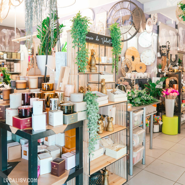 Un espace dédié à la décoration avec des bougies parfumées, des statuettes dorées, des pendules murales et des objets en bois. Un écriteau indique "Envie de Valentin", suggérant une sélection spéciale pour les cadeaux. Les produits sont mis en valeur par des étagères en bois et des plantes suspendues, créant une atmosphère élégante et raffinée.