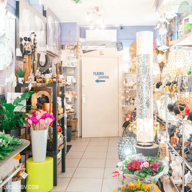Une allée lumineuse du magasin bordée d’étagères contenant des articles de décoration variés : sculptures, cadres, lampes et vases. Au fond, une porte beige avec l’inscription "Fleurs coupées" indique l’accès à une salle réservée aux bouquets de fleurs fraîches.