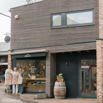 Une boutique d’épicerie fine au design moderne avec un revêtement en bois sombre et une enseigne élégante. Trois femmes souriantes portant des tabliers en toile posent devant l’entrée, donnant une ambiance chaleureuse et artisanale. Un tonneau en bois décoré de pots de fleurs est placé à l’extérieur, renforçant l’esthétique rustique et authentique du lieu.