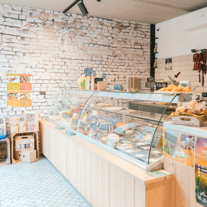 Un comptoir de fromages et de charcuteries artisanales sous une grande vitrine réfrigérée en verre. Le mur en briques blanches donne une touche rustique et moderne à l’espace. Des produits du terroir, des saucissons suspendus et des pâtisseries artisanales sont mis en avant, créant une atmosphère gourmande et authentique.