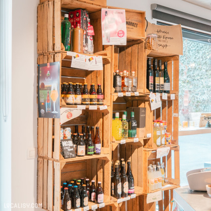Une sélection de boissons locales présentée dans des étagères en bois rustique. Des bières artisanales, des jus naturels et des vins régionaux sont soigneusement disposés avec des étiquettes informatives. L’atmosphère chaleureuse et authentique met en valeur la diversité des produits disponibles.