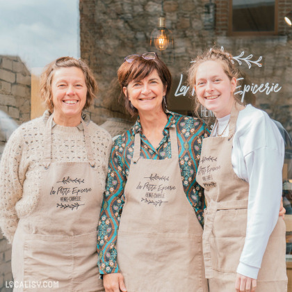 Trois femmes souriantes posent devant la boutique "La Petite Épicerie", vêtues de tabliers beiges brodés du nom de l’établissement. Leur complicité et leur accueil chaleureux transparaissent sur la photo, mettant en avant l’esprit familial et convivial du commerce.