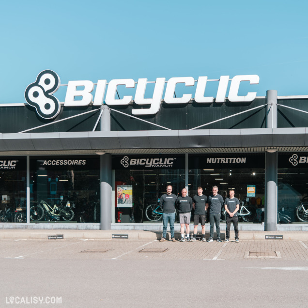 Le magasin Bicyclic avec une grande enseigne blanche et noire sur fond bleu clair. Un groupe de cinq employés en t-shirts noirs posent devant l’entrée du magasin. À travers les grandes vitres, on aperçoit des vélos exposés ainsi que des rayons d’accessoires et de nutrition pour cyclistes.