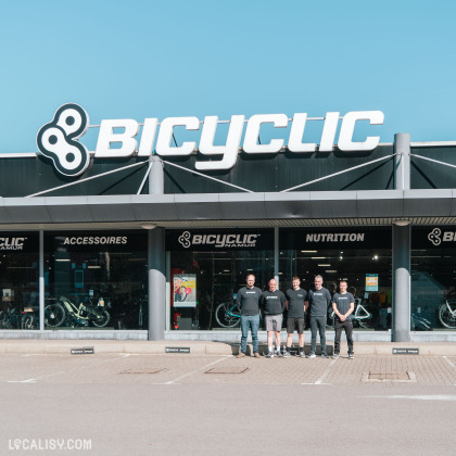 Le magasin Bicyclic avec une grande enseigne blanche et noire sur fond bleu clair. Un groupe de cinq employés en t-shirts noirs posent devant l’entrée du magasin. À travers les grandes vitres, on aperçoit des vélos exposés ainsi que des rayons d’accessoires et de nutrition pour cyclistes.