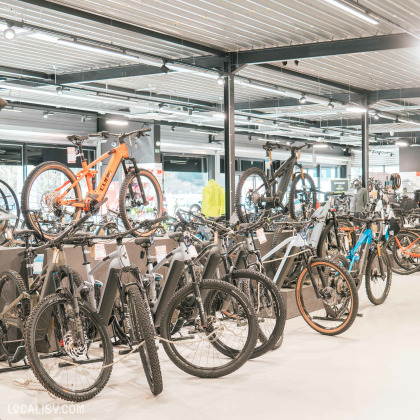 Une section du magasin dédiée aux VTT et vélos électriques, avec des modèles suspendus et d’autres alignés au sol. Un VTT orange Cube est mis en avant sur un présentoir surélevé. L’espace est bien structuré, mettant en valeur la diversité des modèles disponibles.