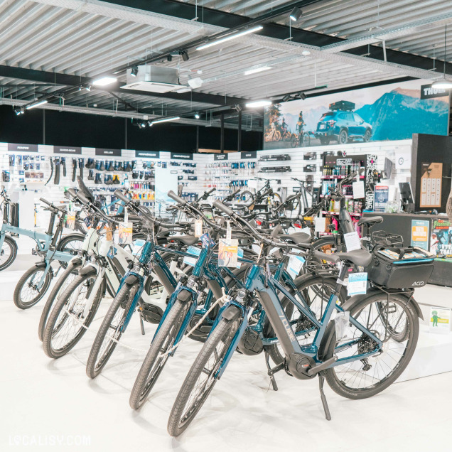 Une section du magasin mettant en avant des vélos électriques urbains alignés en rangées. À l’arrière, un mur d’accessoires comprenant des sacoches, des supports et des équipements de sécurité. Une grande affiche représentant un cycliste en pleine nature renforce l’ambiance sportive et aventureuse du magasin.