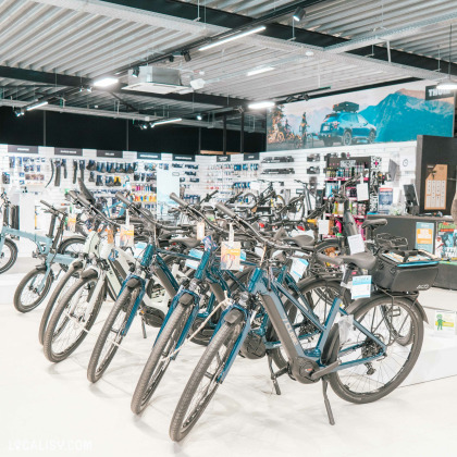 Une section du magasin mettant en avant des vélos électriques urbains alignés en rangées. À l’arrière, un mur d’accessoires comprenant des sacoches, des supports et des équipements de sécurité. Une grande affiche représentant un cycliste en pleine nature renforce l’ambiance sportive et aventureuse du magasin.
