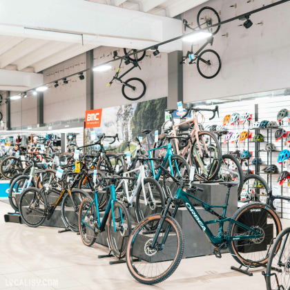 Un vaste espace d’exposition présentant une large sélection de vélos gravel et route alignés sur des supports. Des modèles suspendus au mur apportent une touche visuelle impressionnante. Une variété de casques colorés est disposée en arrière-plan, illustrant la diversité des équipements disponibles.