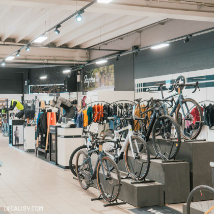 Un rayon dédié aux vêtements et accessoires pour cyclistes, avec des présentoirs soignés et une large gamme de produits. Des vélos haut de gamme sont exposés sur des podiums, mettant en valeur les modèles phares. La marque Rapha est mise en avant sur une bannière murale, soulignant l’accent mis sur les équipements techniques.