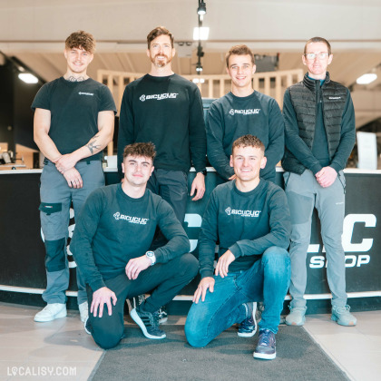 Une équipe de six hommes posant ensemble dans un magasin de vélos. Ils portent des vêtements noirs avec le logo "Bicyclic", certains en sweat-shirts et d'autres en t-shirts. Deux d'entre eux sont accroupis à l'avant, tandis que les quatre autres sont debout derrière un comptoir noir marqué "BICYC... SHOP". L'environnement est lumineux avec un intérieur moderne et du bois en arrière-plan. L'équipe affiche des sourires et une attitude accueillante