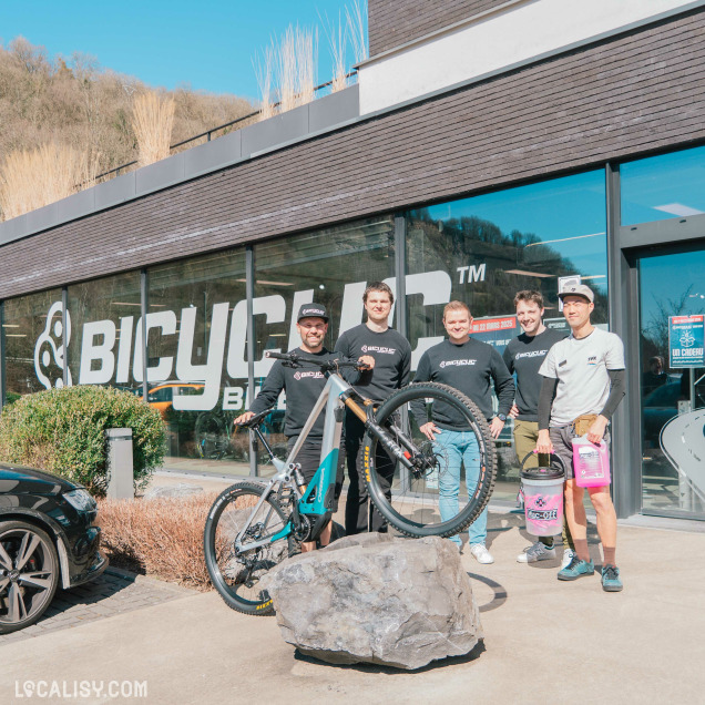 Photo d’équipe devant le Bicyclic Bike Shop Remouchamps, cinq employés posent fièrement avec un vélo de descente sur un rocher, à l’extérieur du magasin vitré en plein soleil.