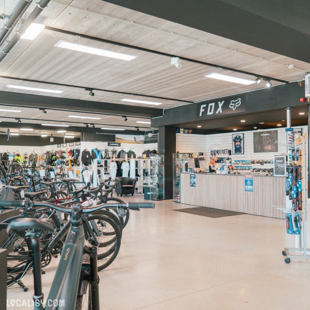 Zone d’accueil et de caisse du Bicyclic Bike Shop Remouchamps, avec des rangées de vélos alignés, des vêtements de sport accrochés aux murs et un comptoir de service sous l’enseigne FOX.