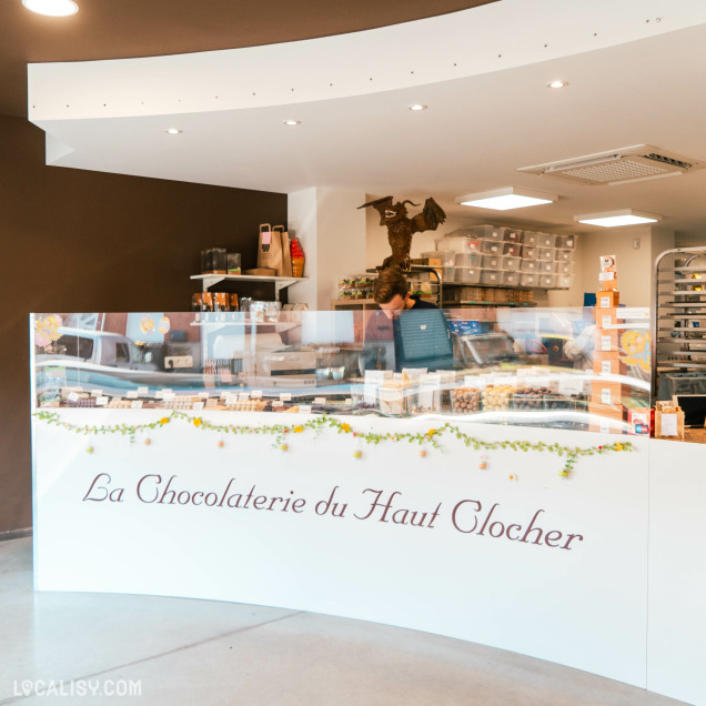 Vue d’ensemble de l’intérieur de la boutique "La Chocolaterie du Haut Clocher" avec le comptoir de vente décoré et l’artisan chocolatier en arrière-plan.