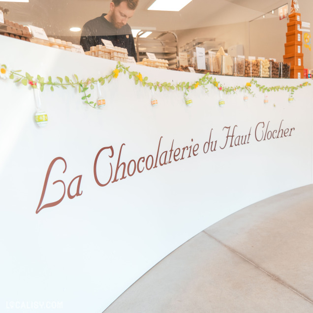 Gros plan sur le nom "La Chocolaterie du Haut Clocher" écrit sur le comptoir blanc décoré avec une guirlande florale.